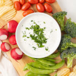 Overhead view of ranch dressing in a bowl sitting on a cutting board with potato chips and raw vegetables.