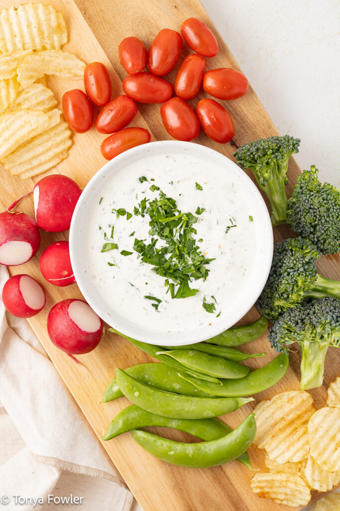 Overhead view of ranch dressing in a bowl sitting on a cutting board with potato chips and raw vegetables.