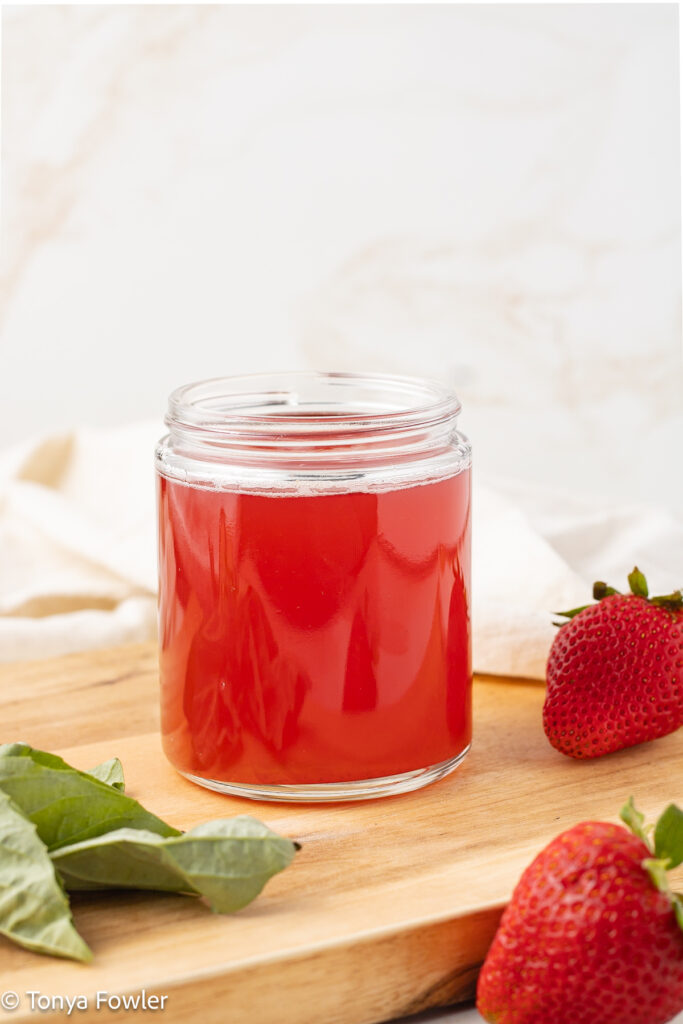 Straight on view of Strawberry Basil Syrup in a jar sitting on a cutting board with fresh berries and basil.