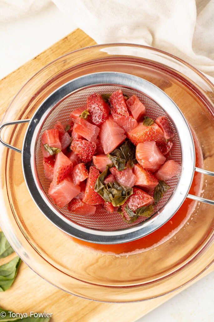 Cooked strawberries and basil are being strained into a bowl.