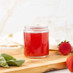Straight on view of fruit syrup in a jar sitting on a cutting board with fresh strawberries and basil near by.