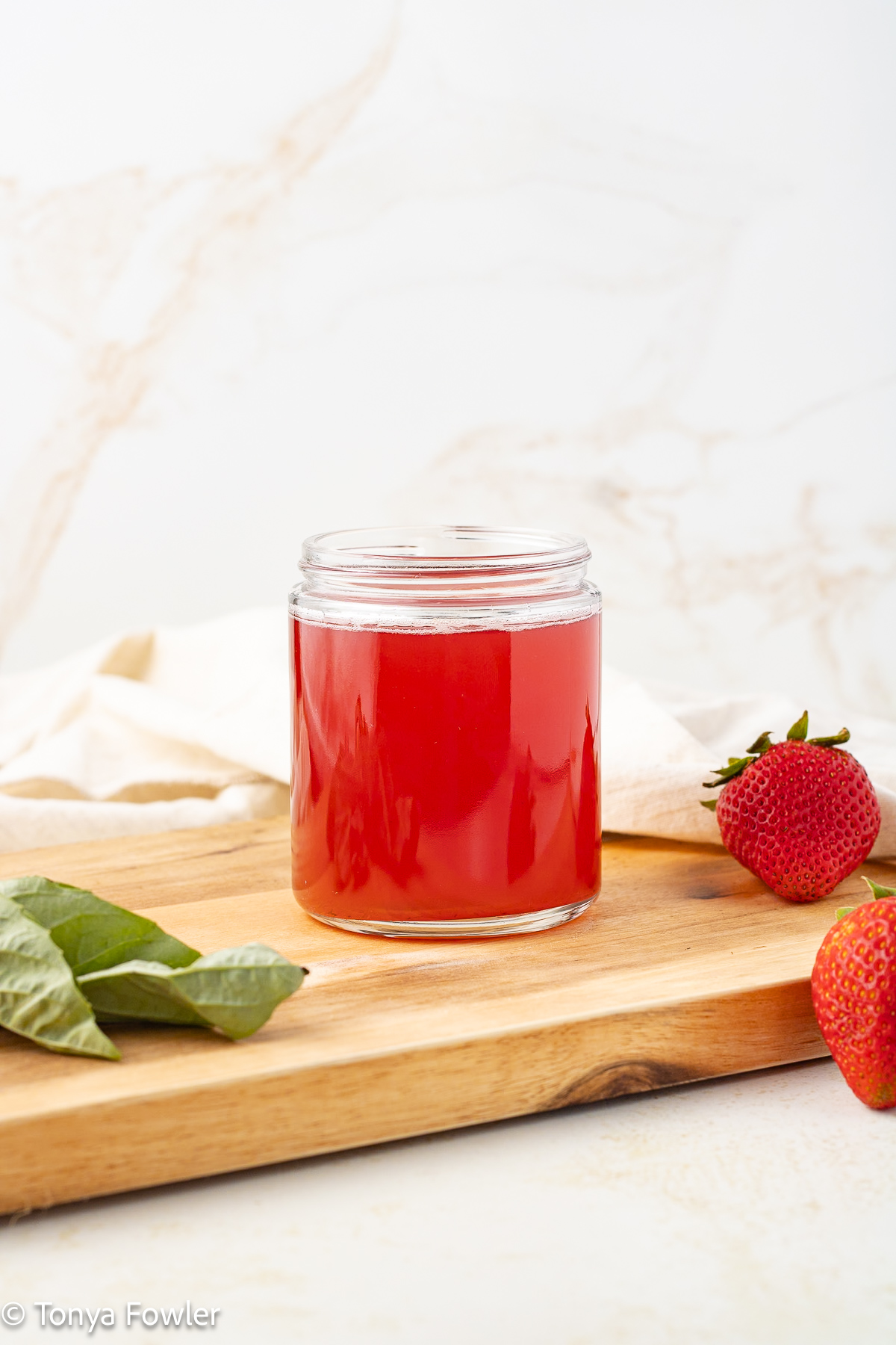 Straight on view of fruit syrup in a jar sitting on a cutting board with fresh strawberries and basil near by.