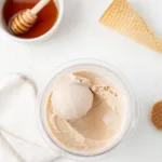 Scoop of honey ice cream in a pint container surrounded by honey and honey wafers.