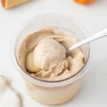 A scoop of pumpkin ice cream in a container with a spoon. Ice cream cones and pumpkins in the background.