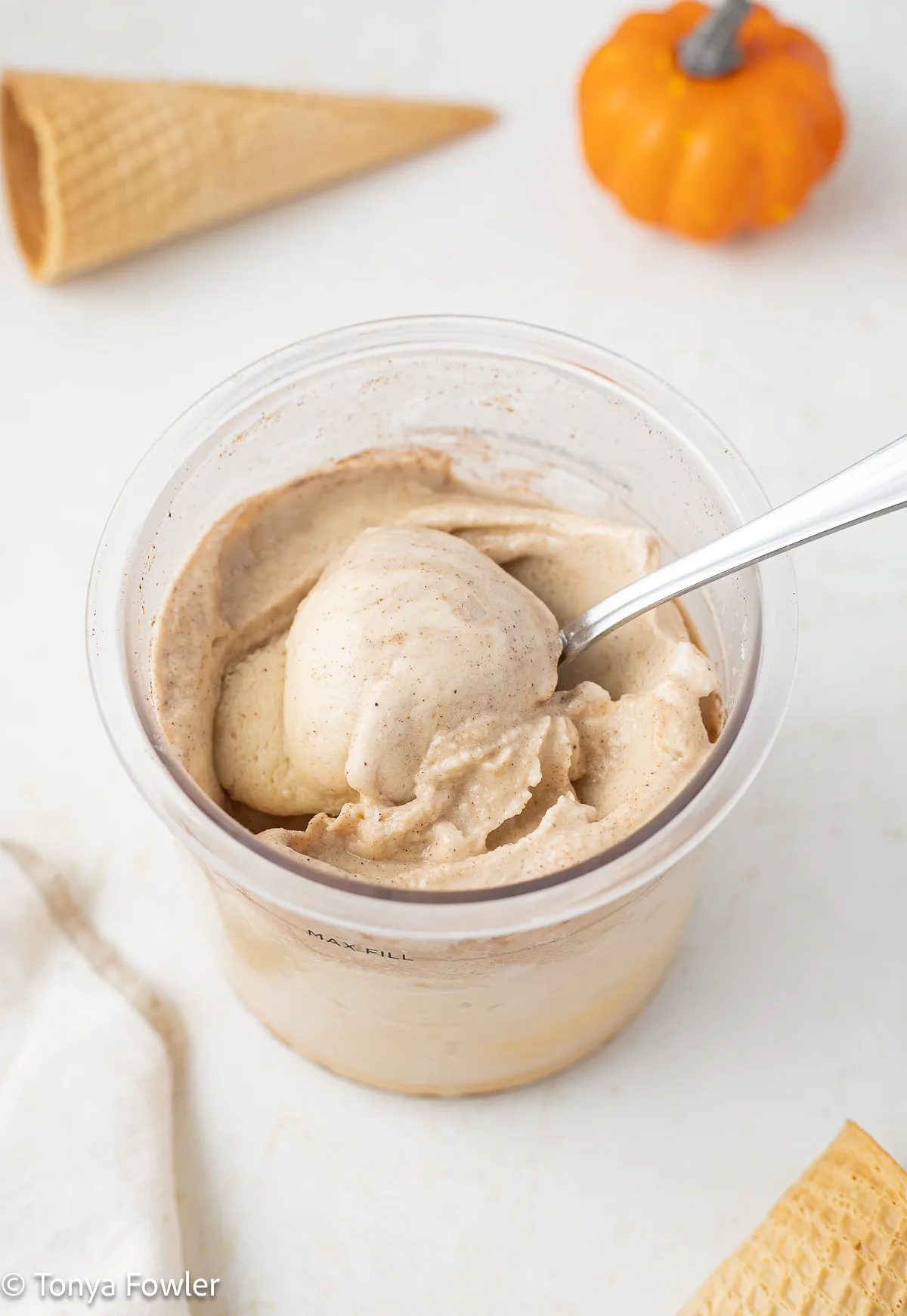 A scoop of pumpkin ice cream in a container with a spoon. Ice cream cones and pumpkins in the background.