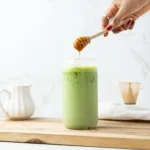 Salted honey matcha latte with a hand holding a honey dipper.
