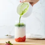 Hand pouring matcha into a glass with milk and strawberries.