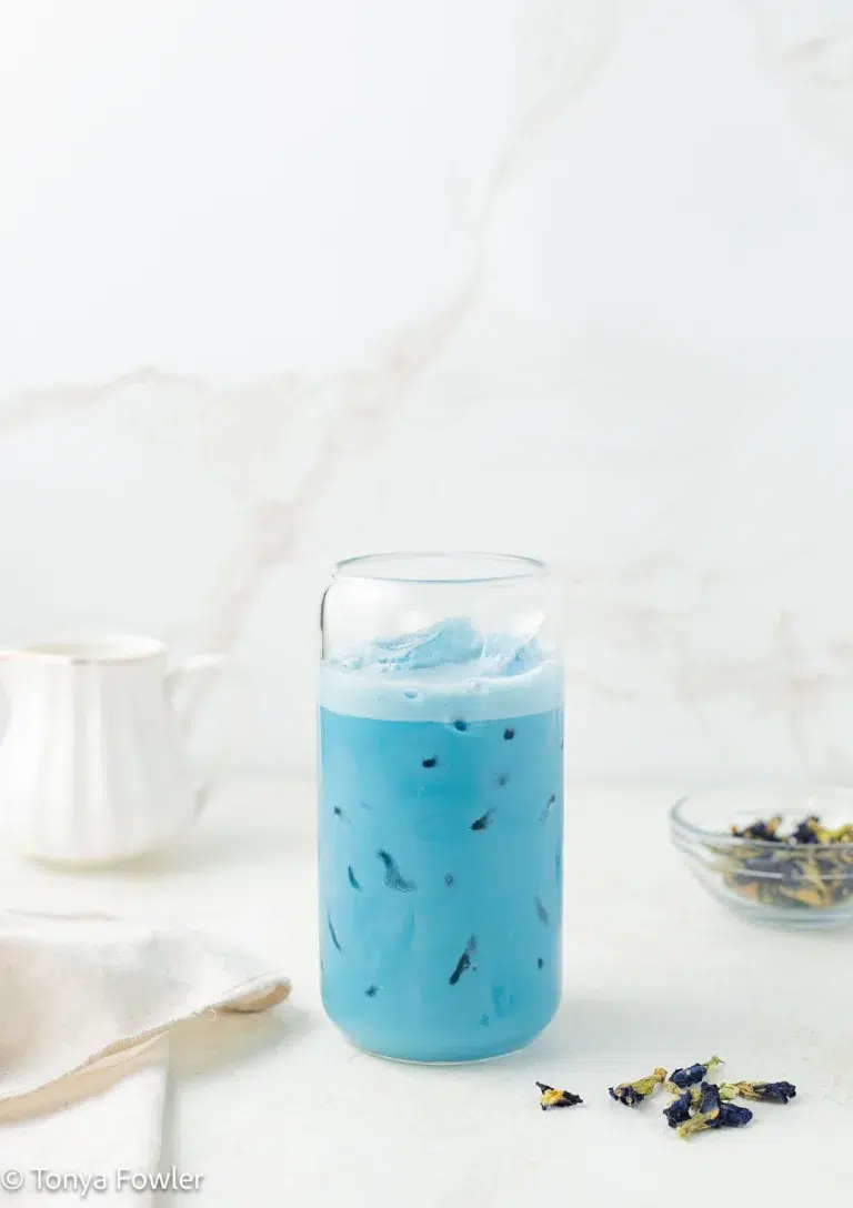 Butterfly pea flower latte in a glass with butterfly pea flower petals on a counter.